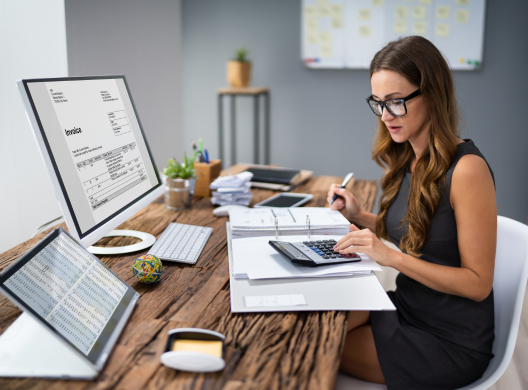 Young woman working at a desk in front of a computer using a calculator. An invoice is on her computer screen.