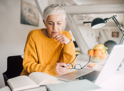 Older woman working at desk with open book and papers and laptop.