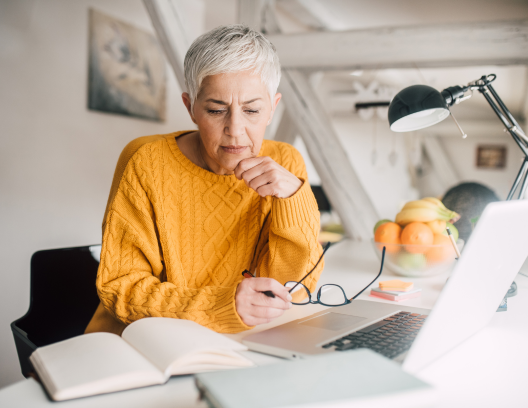 Older woman working at desk with open book and papers and laptop.