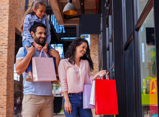 Photo of mother, father, and child. Child is on the father's shoulders. They are shopping together and smiling and carrying shopping bags. They are happy and smilling.