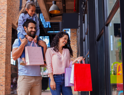 Photo of mother, father, and child. Child is on the father's shoulders. They are shopping together and smiling and carrying shopping bags. They are happy and smilling.